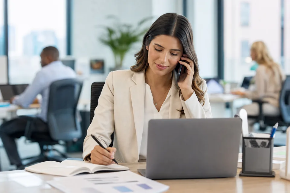Account manager on a client call taking notes at her desk in an open plan office